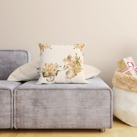 Față de pernă decorativă din bumbac 100%, 45x45cm, cu un iepuraș simpatic care transportă un cărucior plin cu flori de primăvară, pe fundal crem pastelat.
